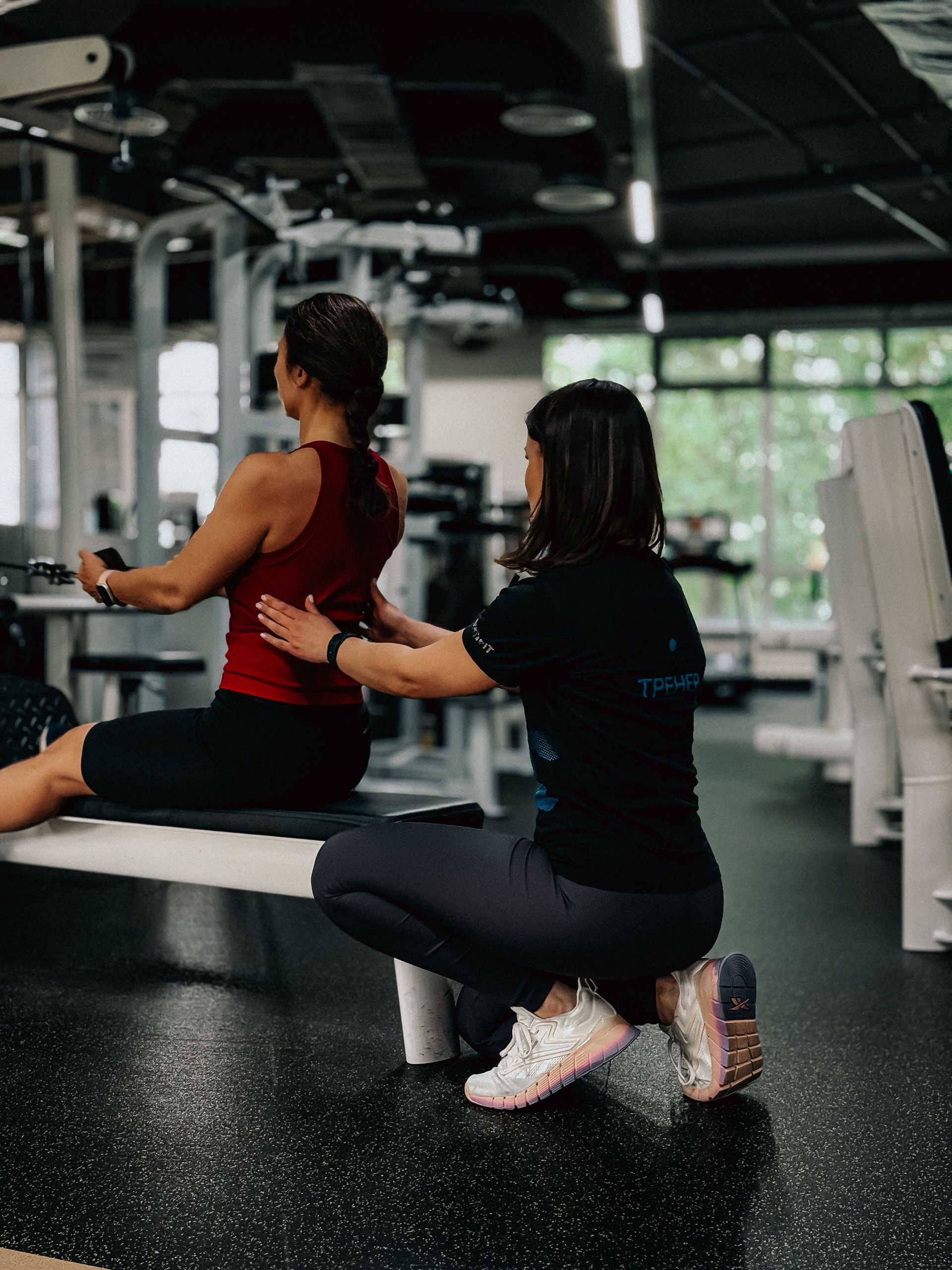 Two people exercising on a gym machine, one seated and one kneeling nearby.