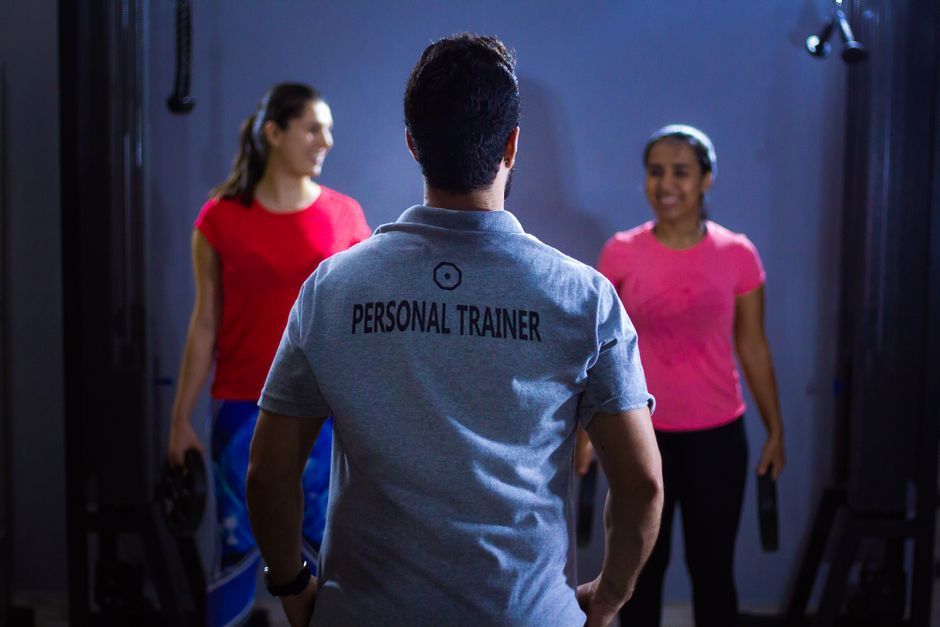 Personal trainer facing two clients in a dim gym, with one woman in red and another in pink.