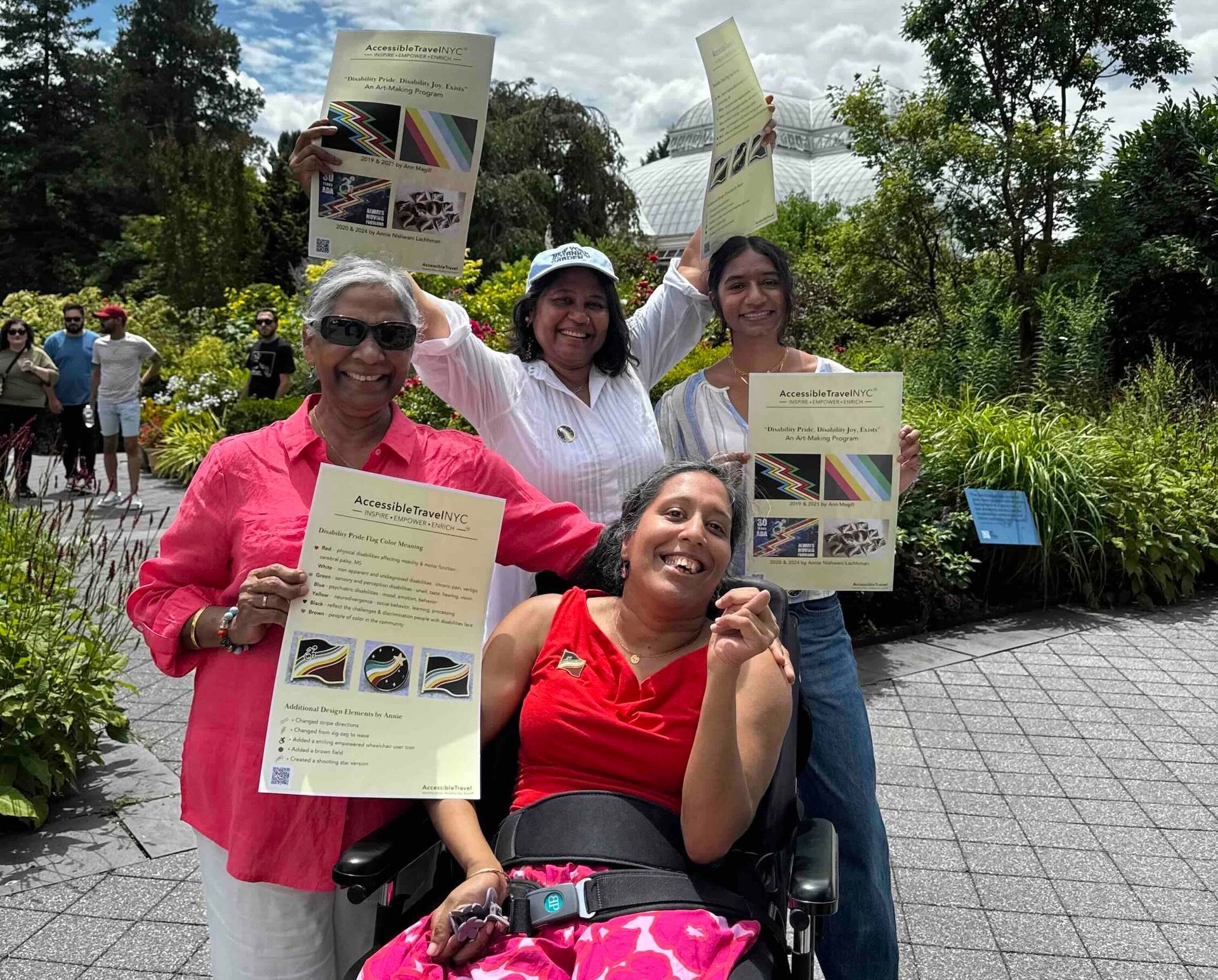 Lakshmee, Annie and family smiling widely at NYBG holding up signs of Annie's Disability Pride flag. 