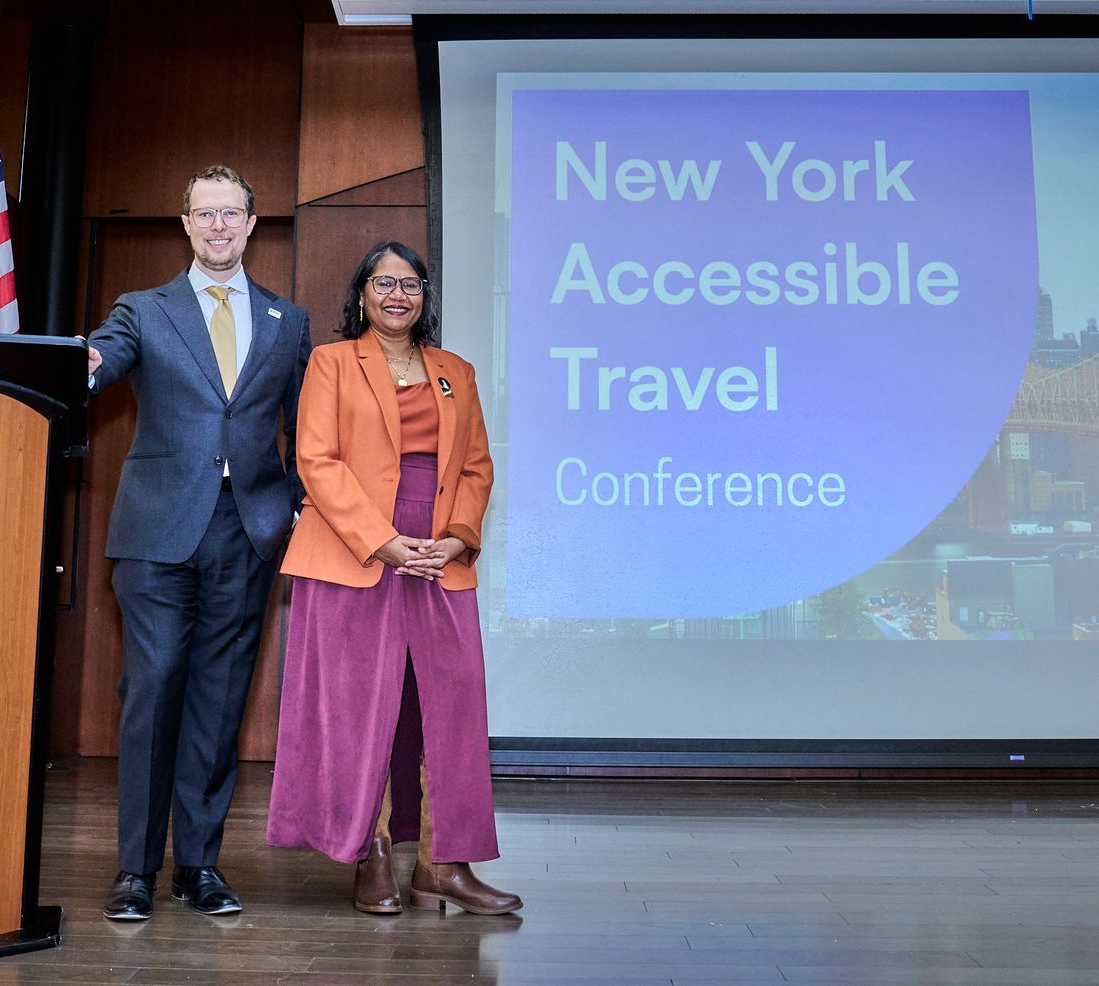 Lakshmee and Ben stands on stage with the New York Accessible Travel Conference graphics behind.