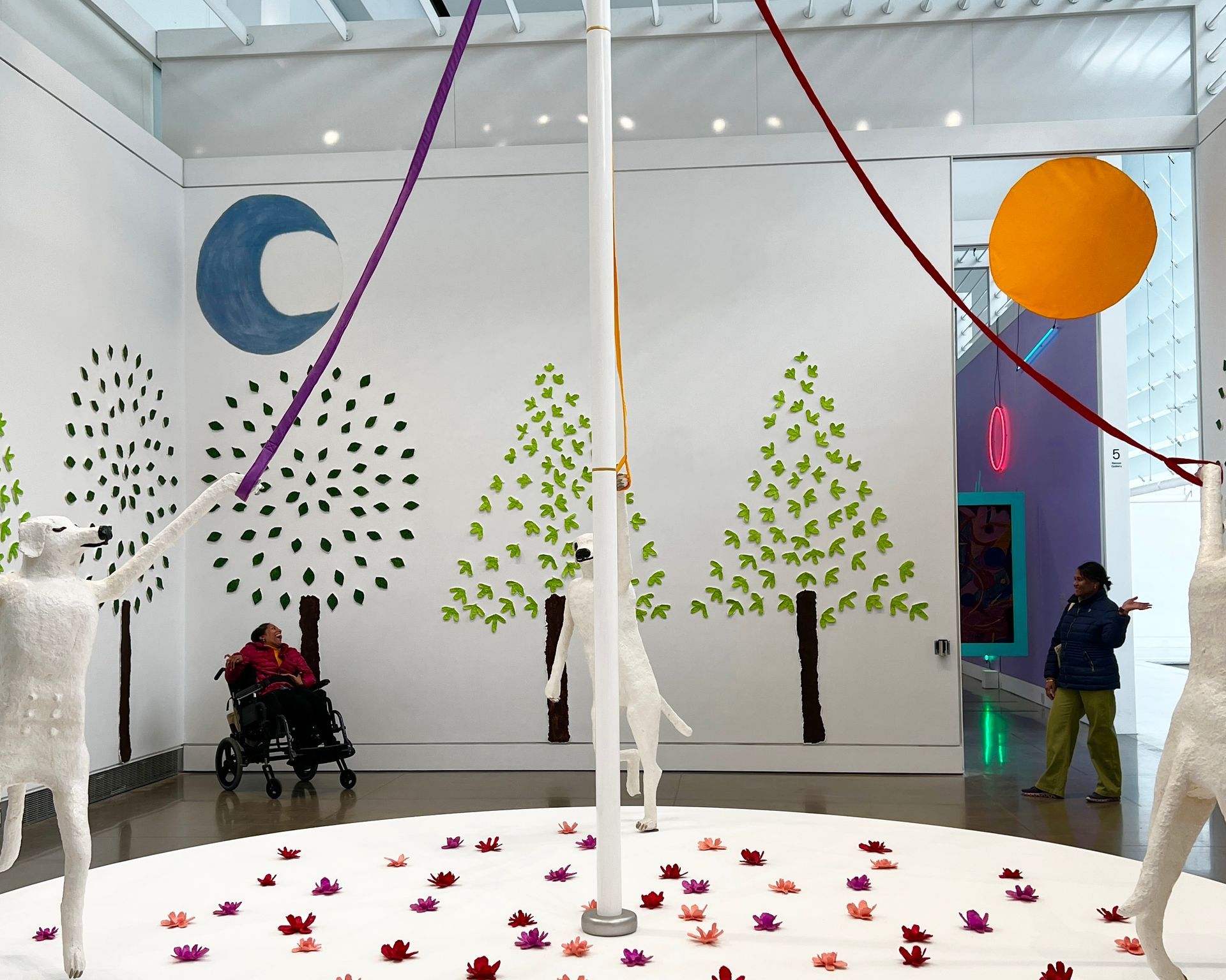 The art installation is in a bright space. Ribbons drape from the ceiling converging on a pole with 3 white dog figures. 