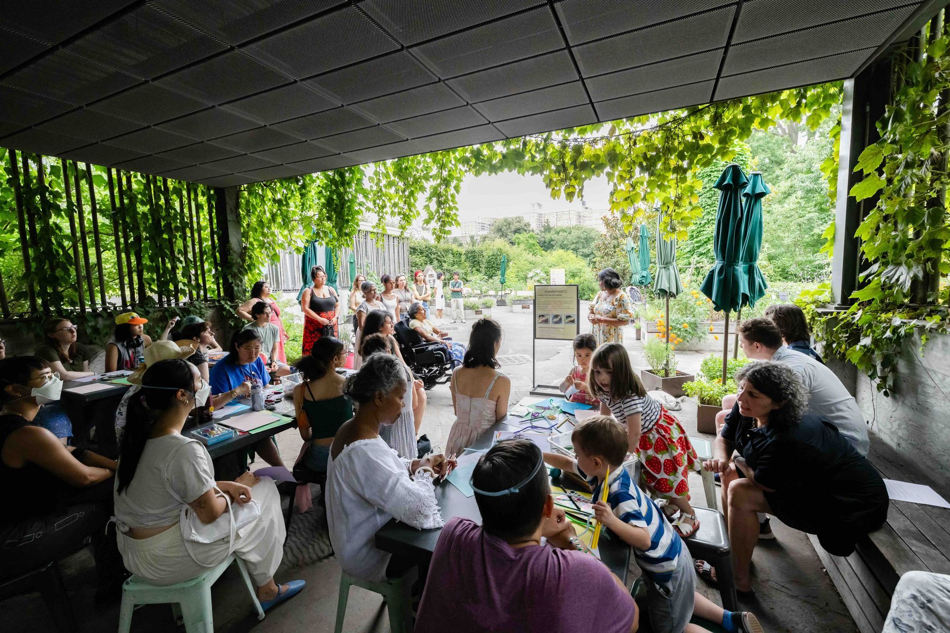A photo taken of the group from the back of the program area showcasing many participants actively listening and creating. Lakshmee and Annie are in front leading the discussion.  The spot is beautifully adorned by lush green plants. 