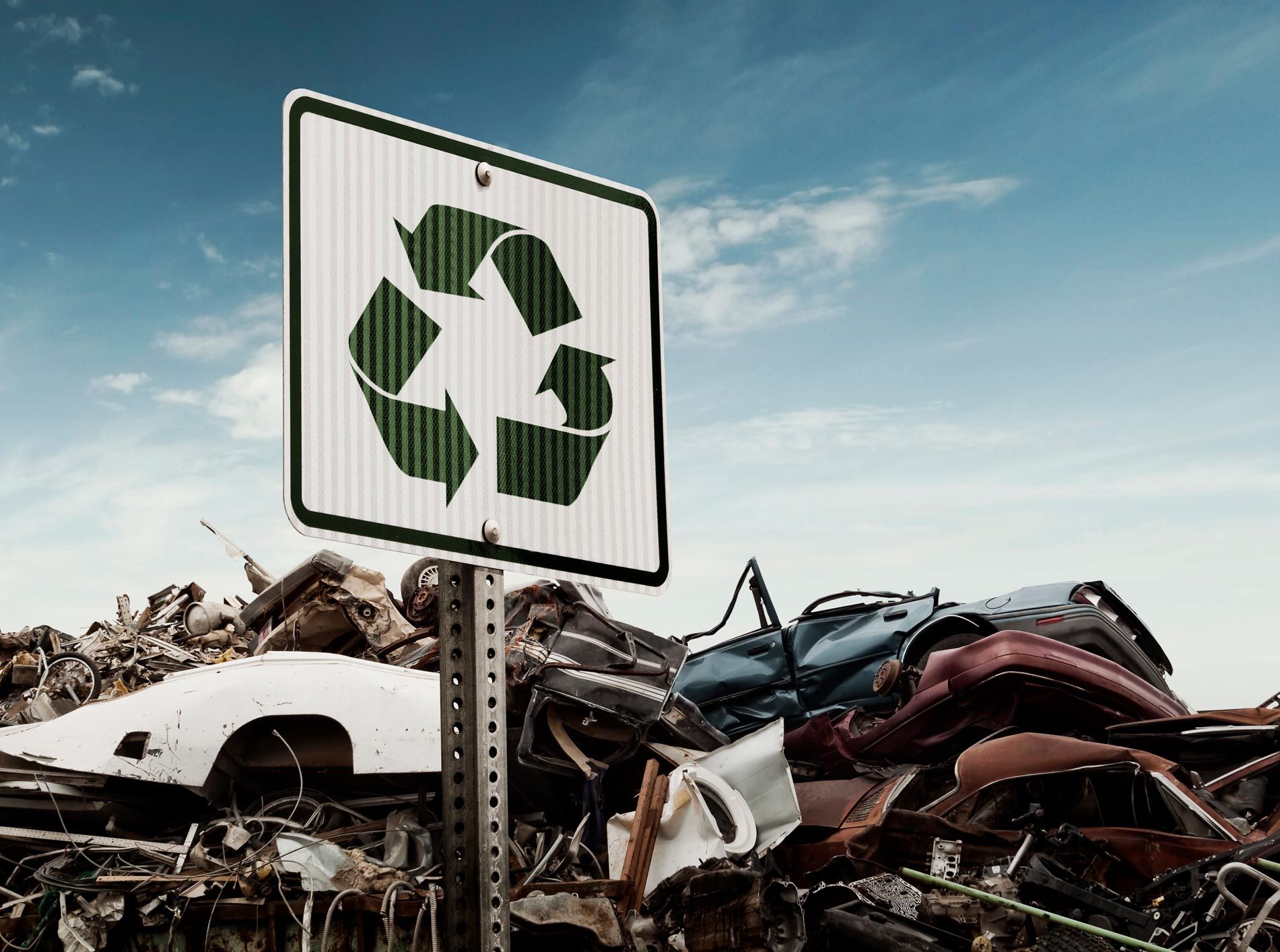 Recycling sign in front of a pile of scrap cars, under a blue sky.
