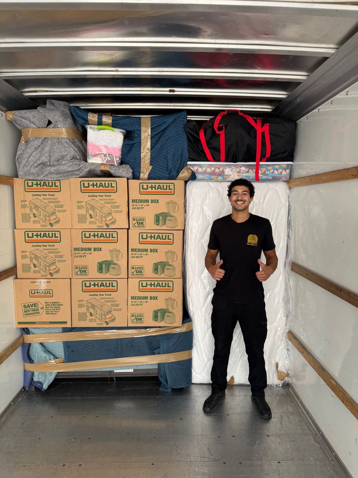 Man in yellow shirt gives thumbs-up, standing on red carpet next to a hand truck carrying a cardboard-covered piece of furniture.