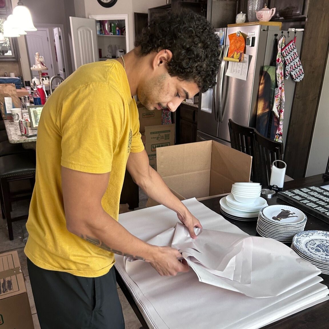 Person in yellow shirt wrapping dishes with paper at a table, preparing for a move.