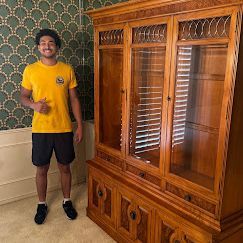 Man in yellow shirt and shorts stands by a large wooden cabinet in front of patterned wallpaper.