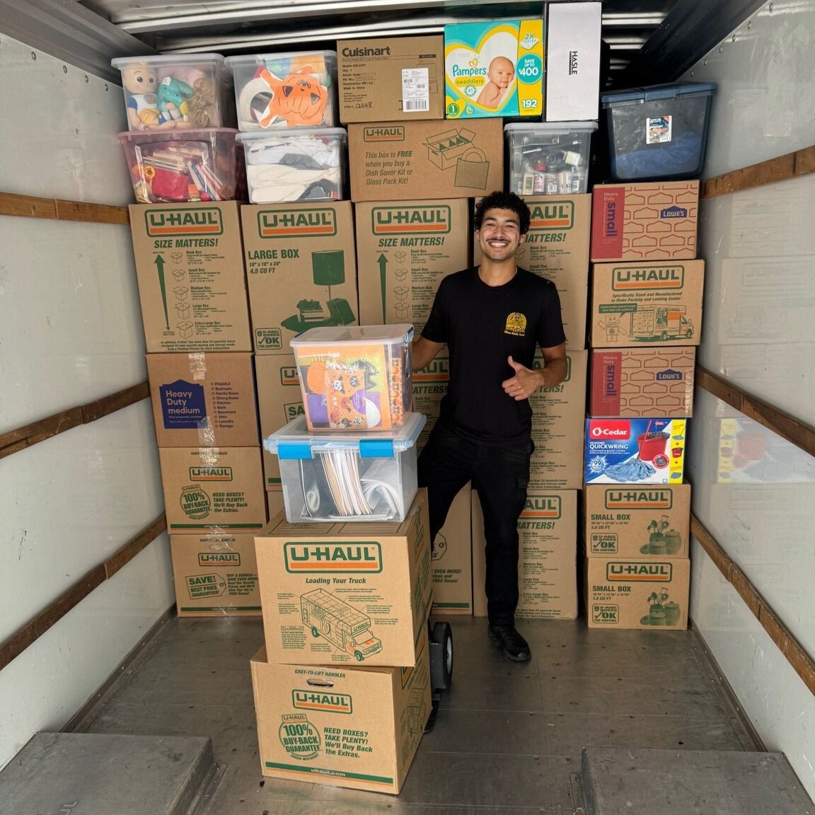 Man in black thumbs-up inside a U-Haul truck, surrounded by stacked moving boxes and bins.