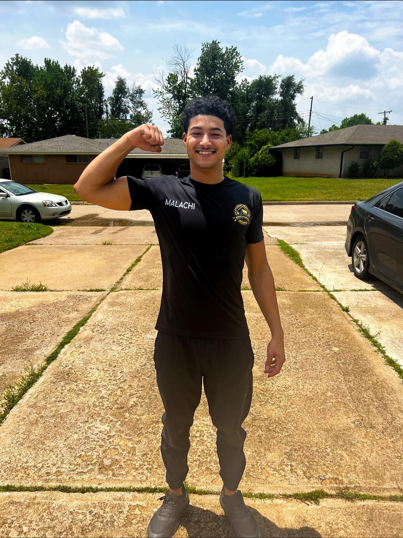 Man flexing arm, smiling, in front of houses and cars.
