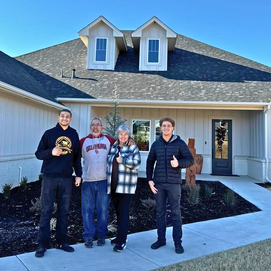 Four people smiling in front of a new house. They are making thumbs-up gestures on a sunny day.
