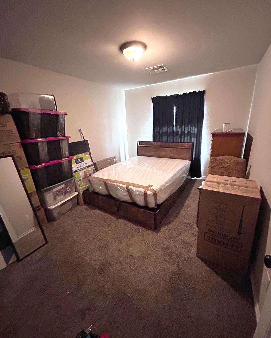 Bedroom with bed, boxes, and storage bins; brown carpet, dark curtains, and a ceiling light.