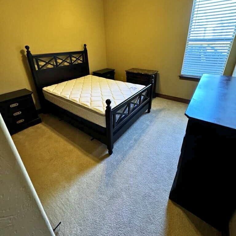 Bedroom with a black bed, nightstands, and dresser. Neutral carpet and wall colors, window with blinds.