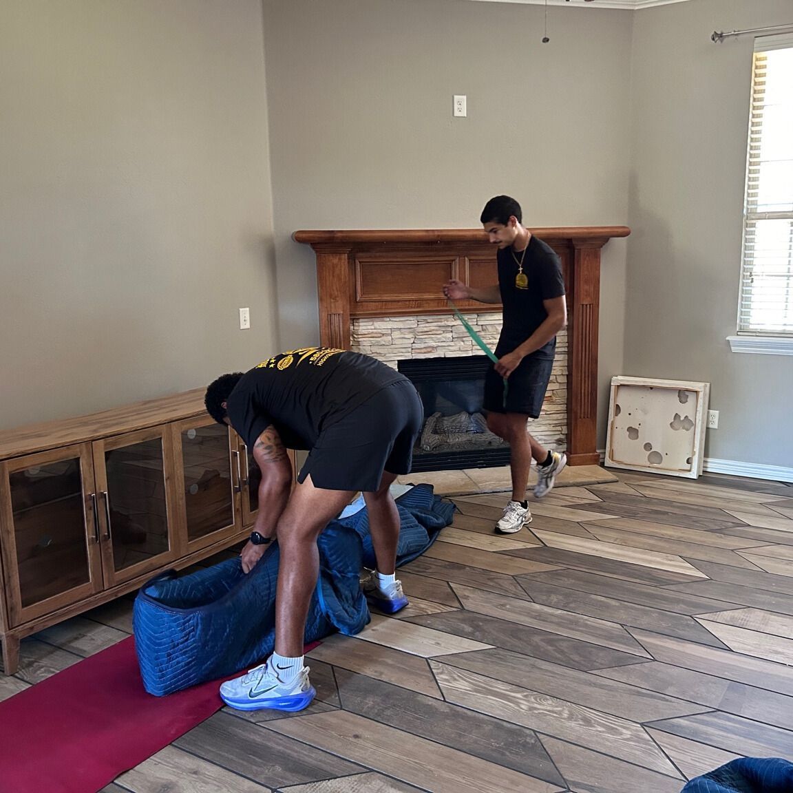 Two people moving furniture in a living room with wooden floors, fireplace, and window.