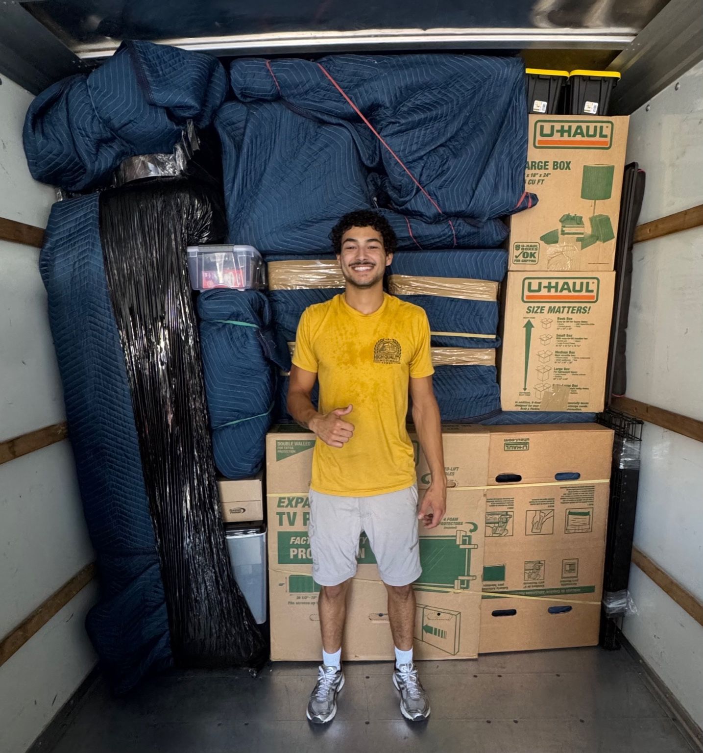 Man in yellow shirt gives thumbs-up, standing on red carpet next to a hand truck carrying a cardboard-covered piece of furniture.