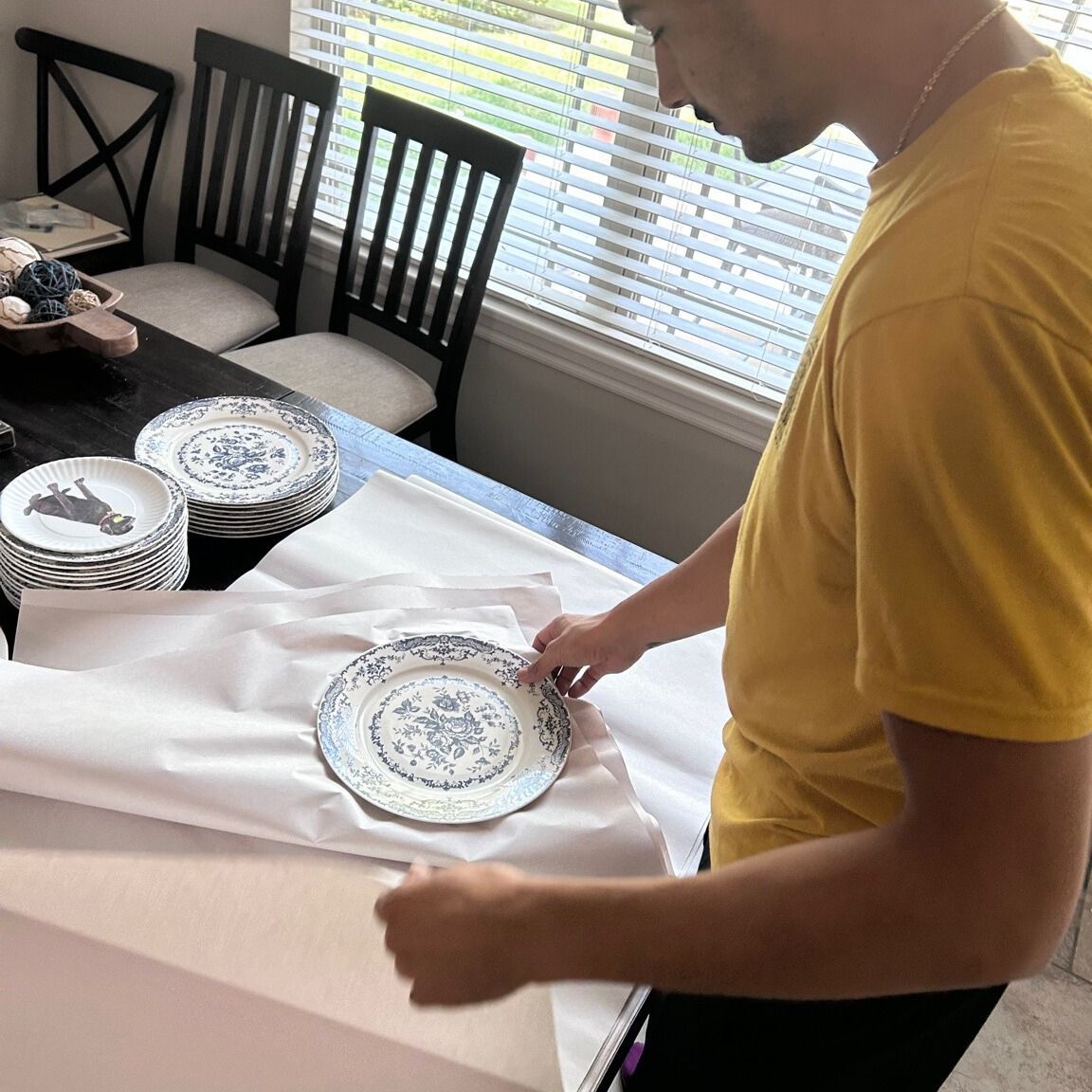 Person wrapping a blue-and-white plate on a table with a stack of other plates.