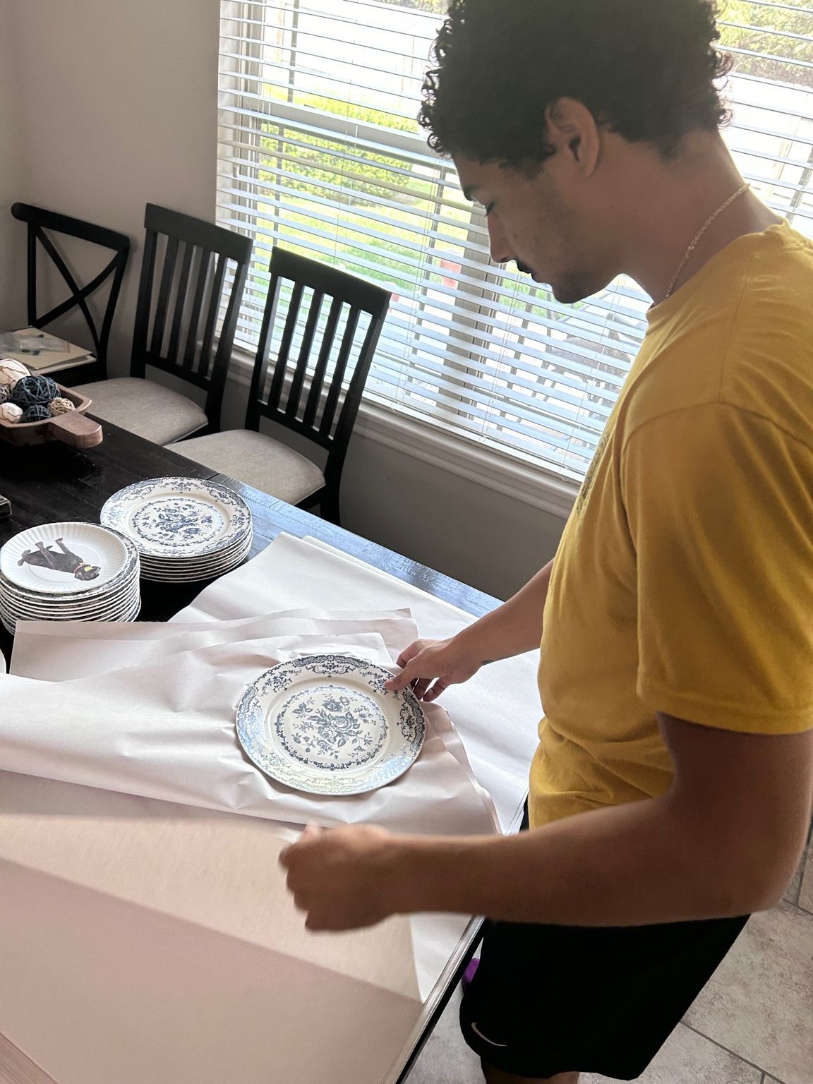 Man wrapping blue and white plate in white paper on a table. Dining table with chairs visible in the background.