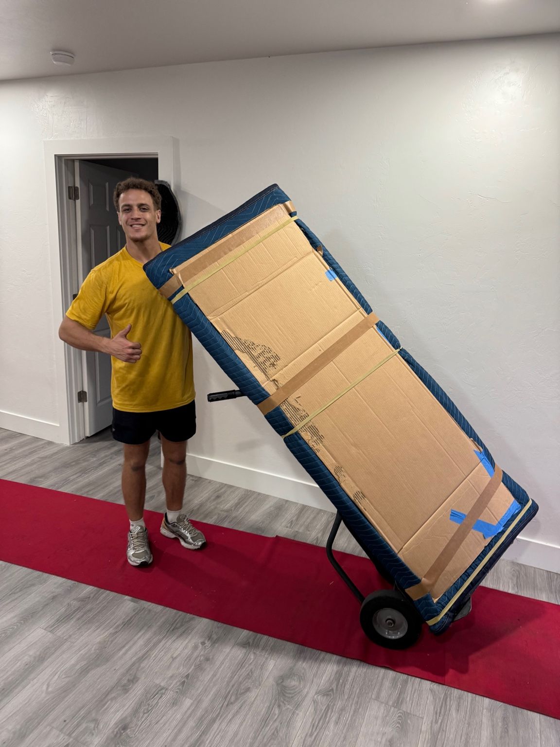 Man in yellow shirt gives thumbs-up, standing on red carpet next to a hand truck carrying a cardboard-covered piece of furniture.