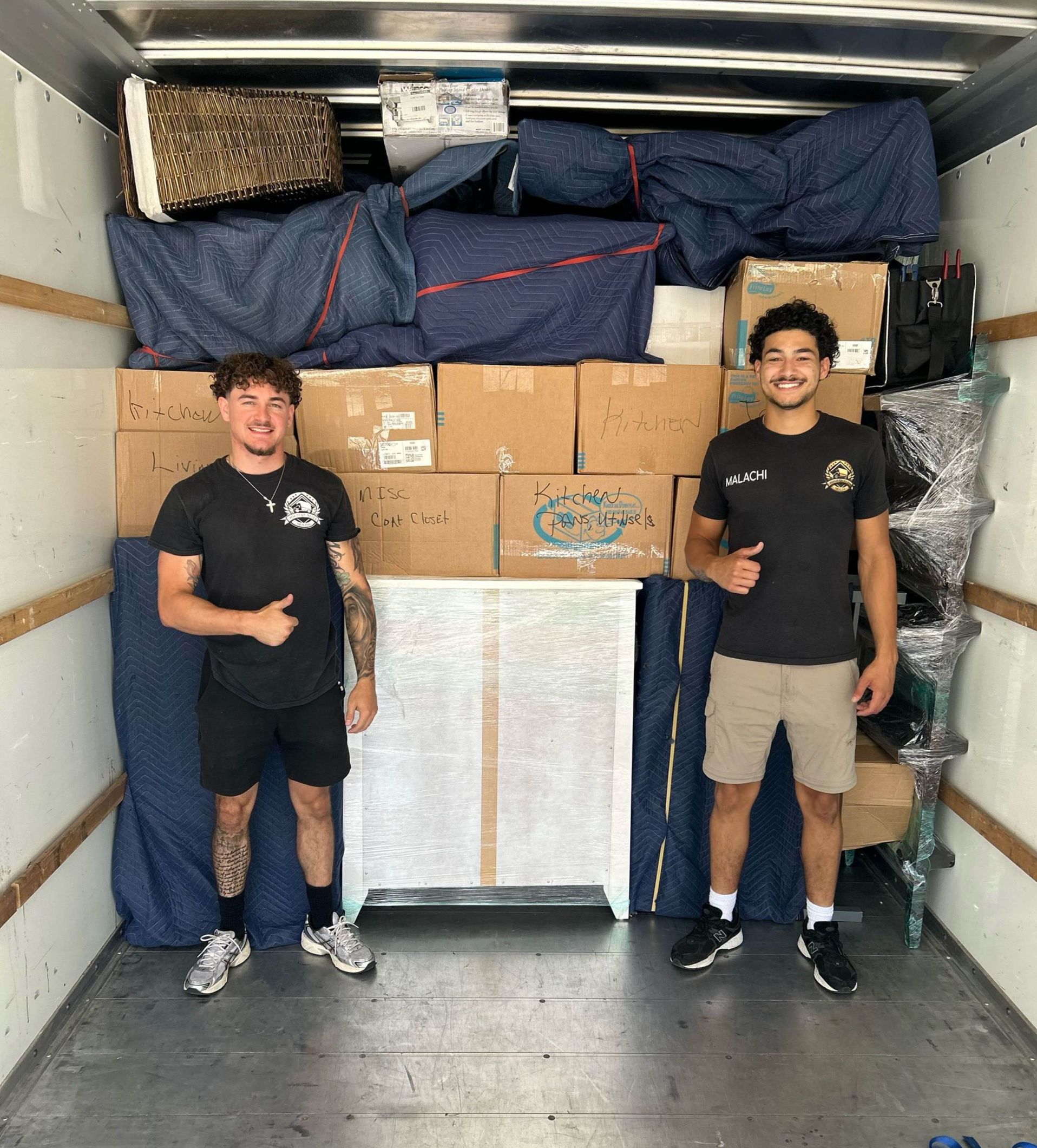 Two men stand in a moving truck filled with packed boxes and furniture. They smile and give thumbs up.