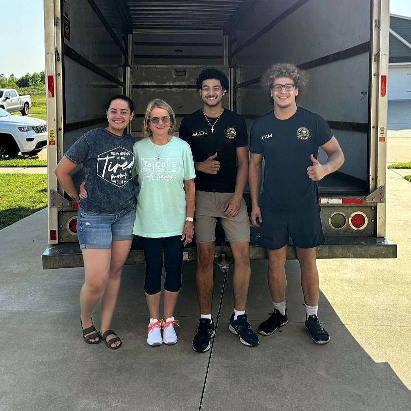 Four people pose in front of a moving truck; two women, two men, thumbs up. Outdoors.