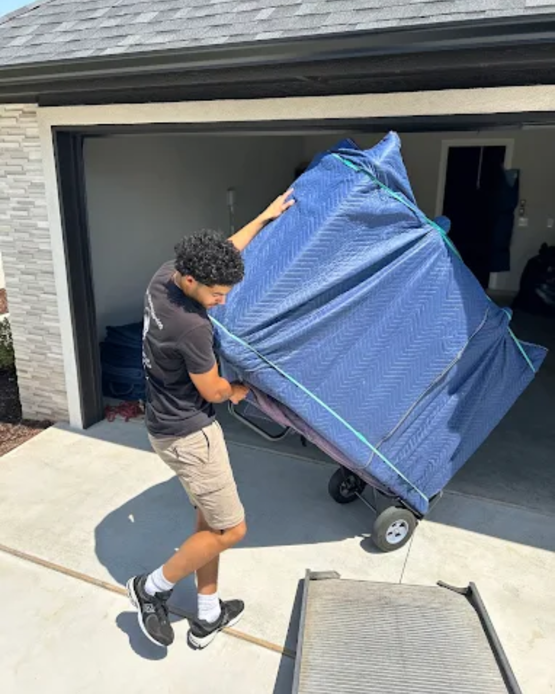 Man moving furniture, wrapped in blue pad, on a hand truck, exiting a garage onto a ramp.