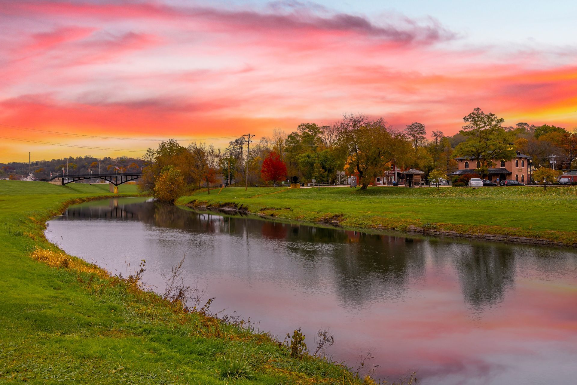 Sunset over a calm river with pink sky, grassy banks, and houses in the distance