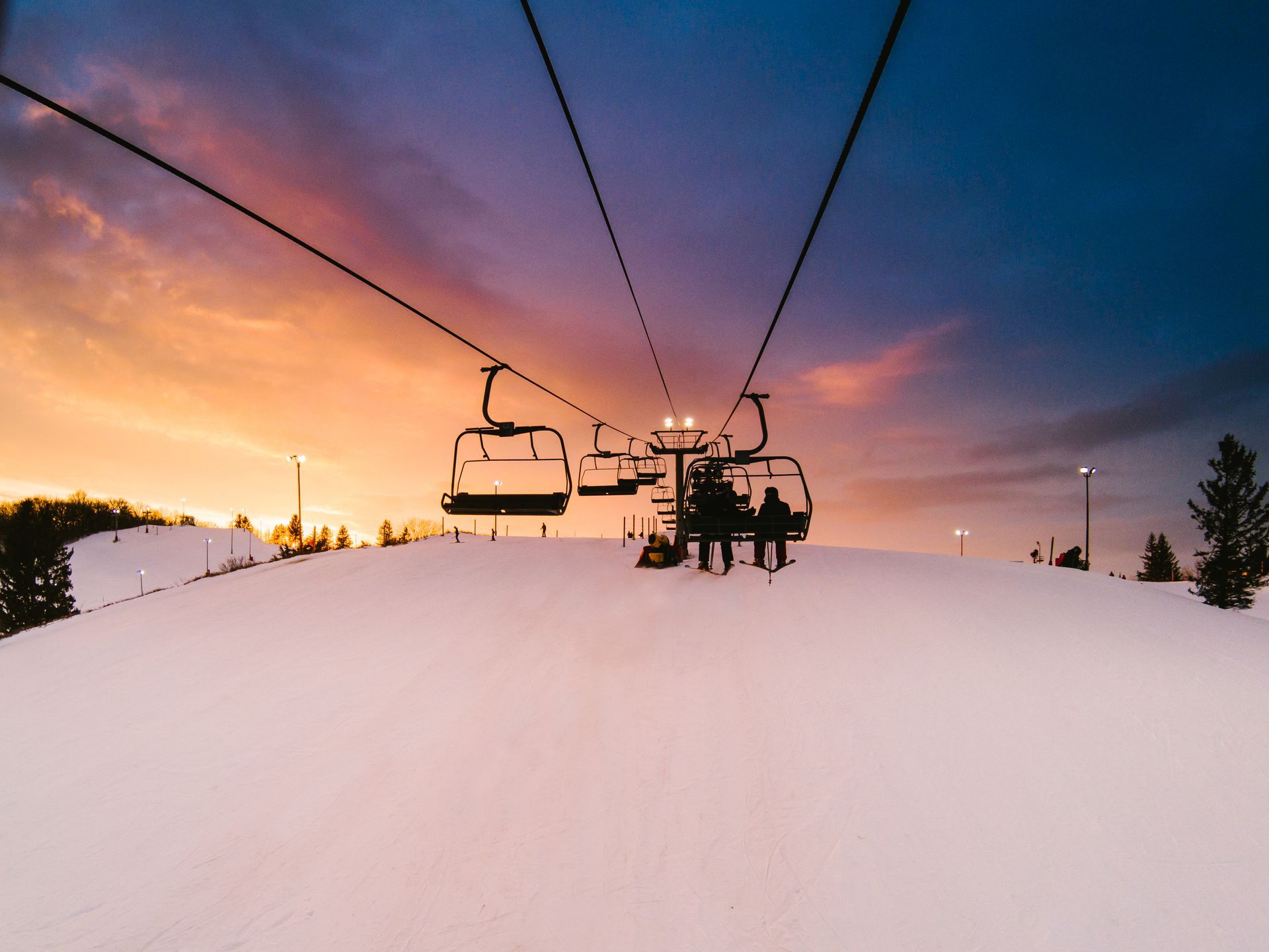 Ski lift chairs ascending a snowy slope at sunset, with pink-orange sky and silhouetted riders