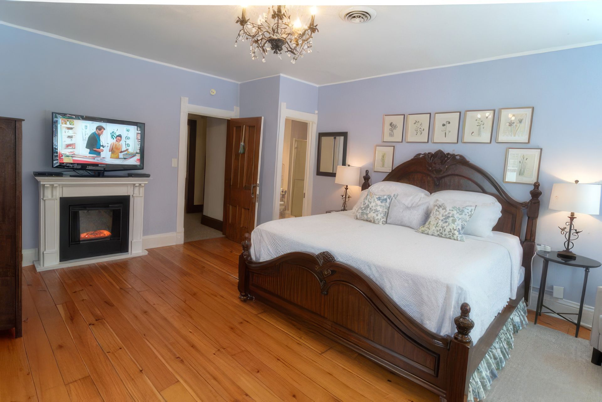 Bedroom with hardwood floors, fireplace, wall-mounted TV, and a large wooden bed with white bedding.