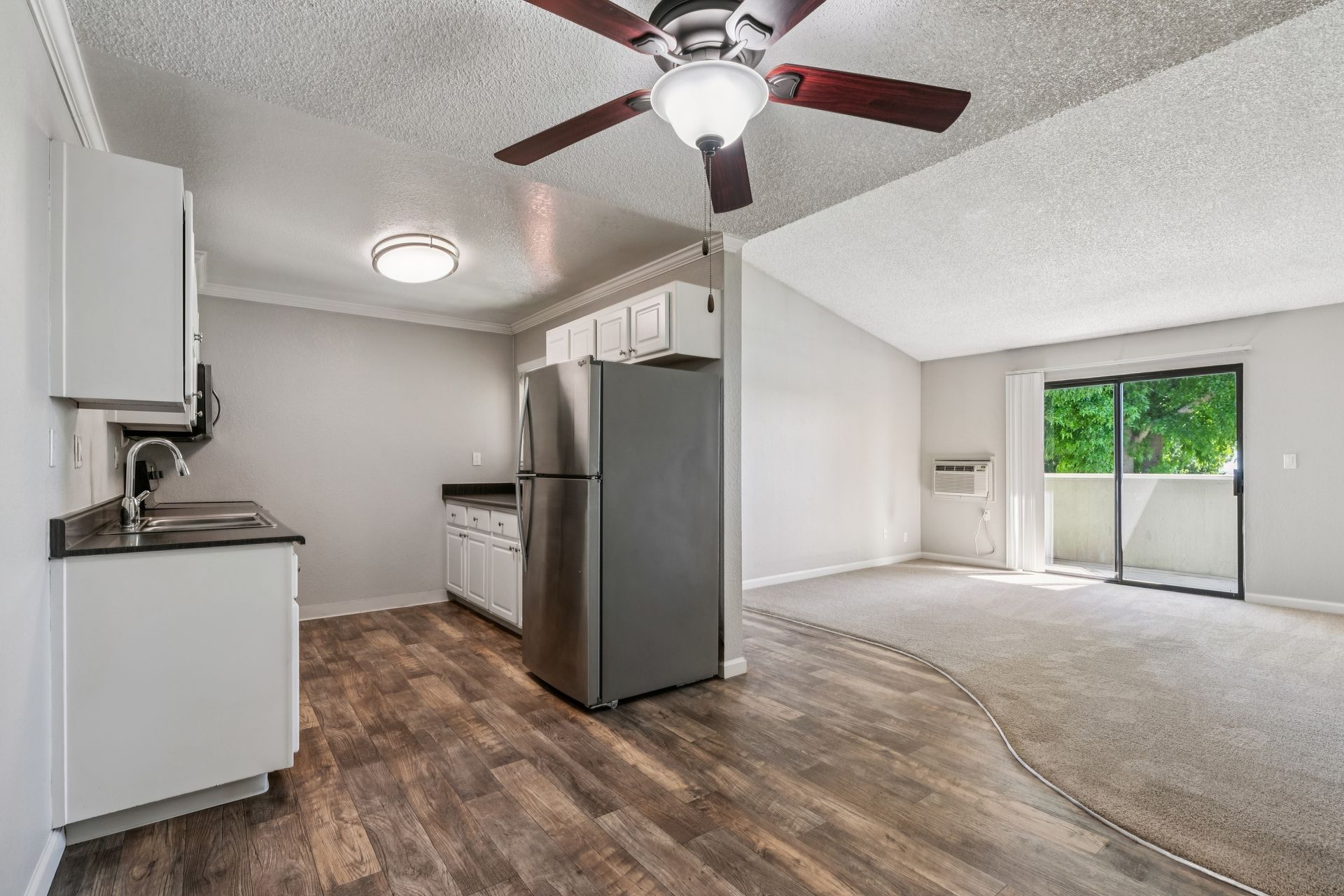 Empty apartment living area with kitchen, ceiling fan, wood floors, and sliding glass door.