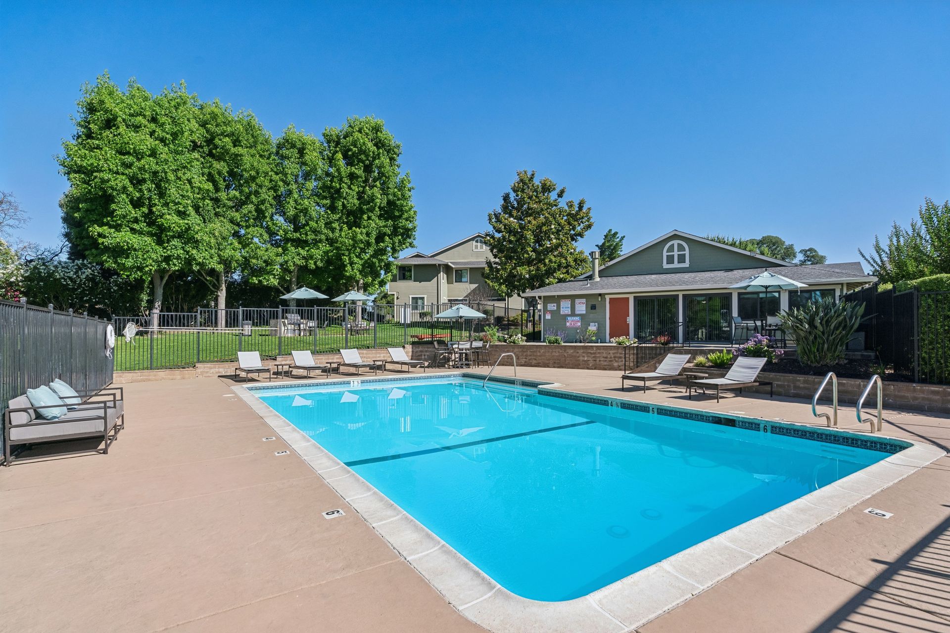 Outdoor swimming pool with blue water, lounge chairs, and trees beside a clubhouse under a clear sky