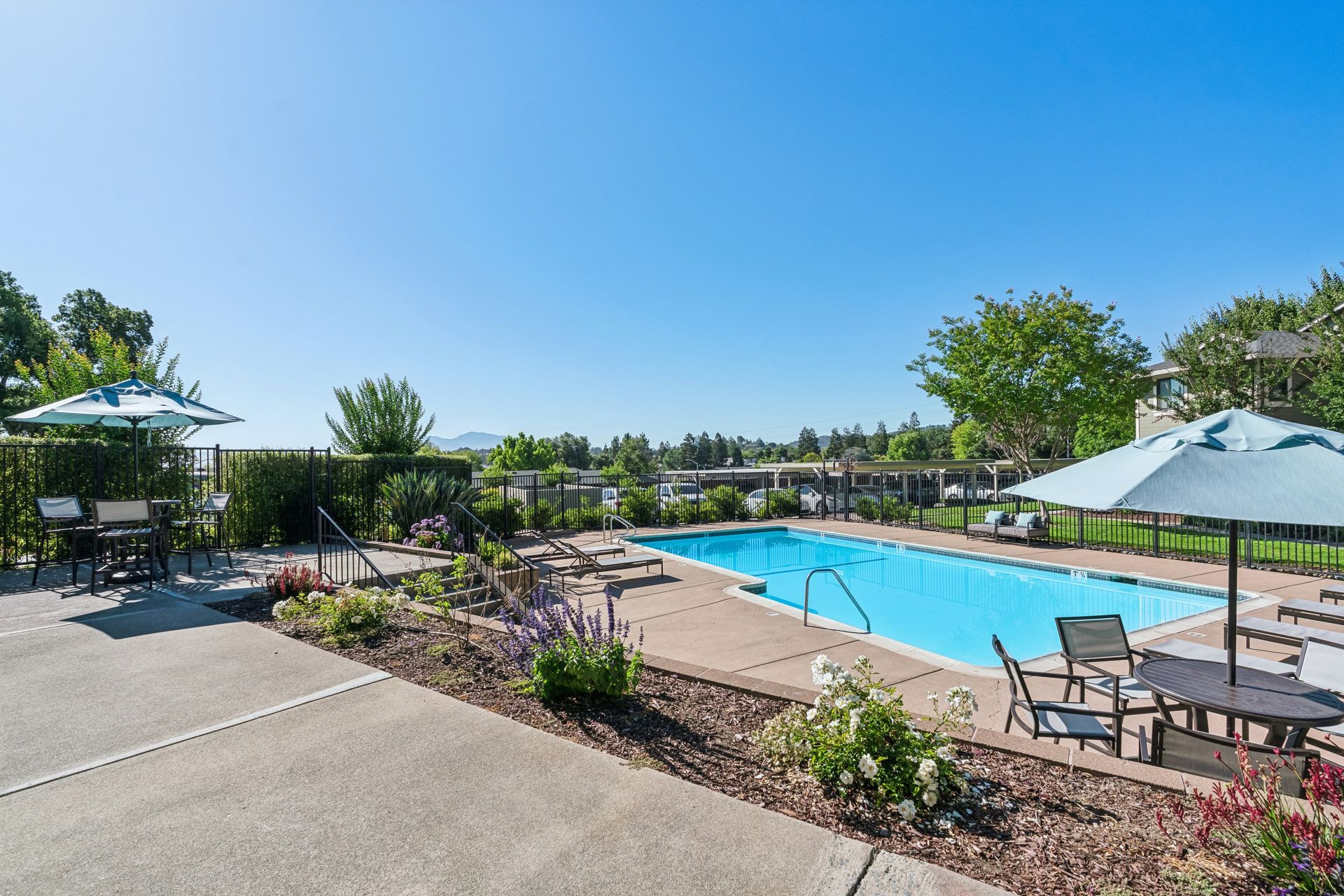 Outdoor swimming pool with lounge chairs, umbrellas, and landscaped patio under a clear blue sky