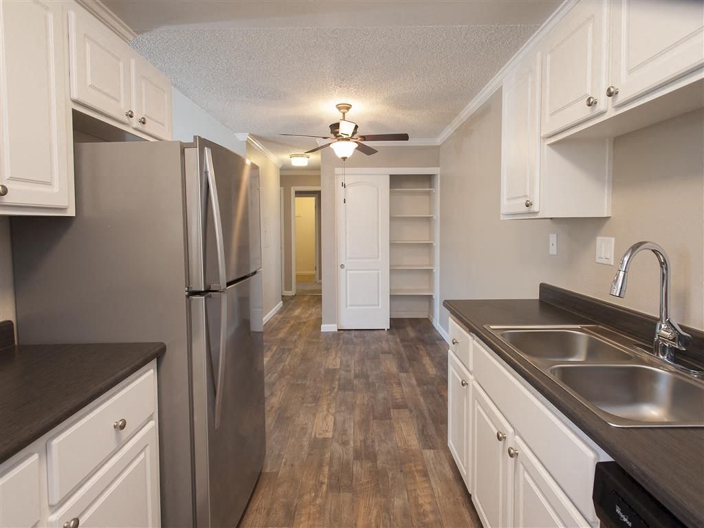 Galley-style apartment kitchen with white cabinets, dark countertops, a stainless steel fridge, and a double sink at Ridgecrest Apartment Homes.