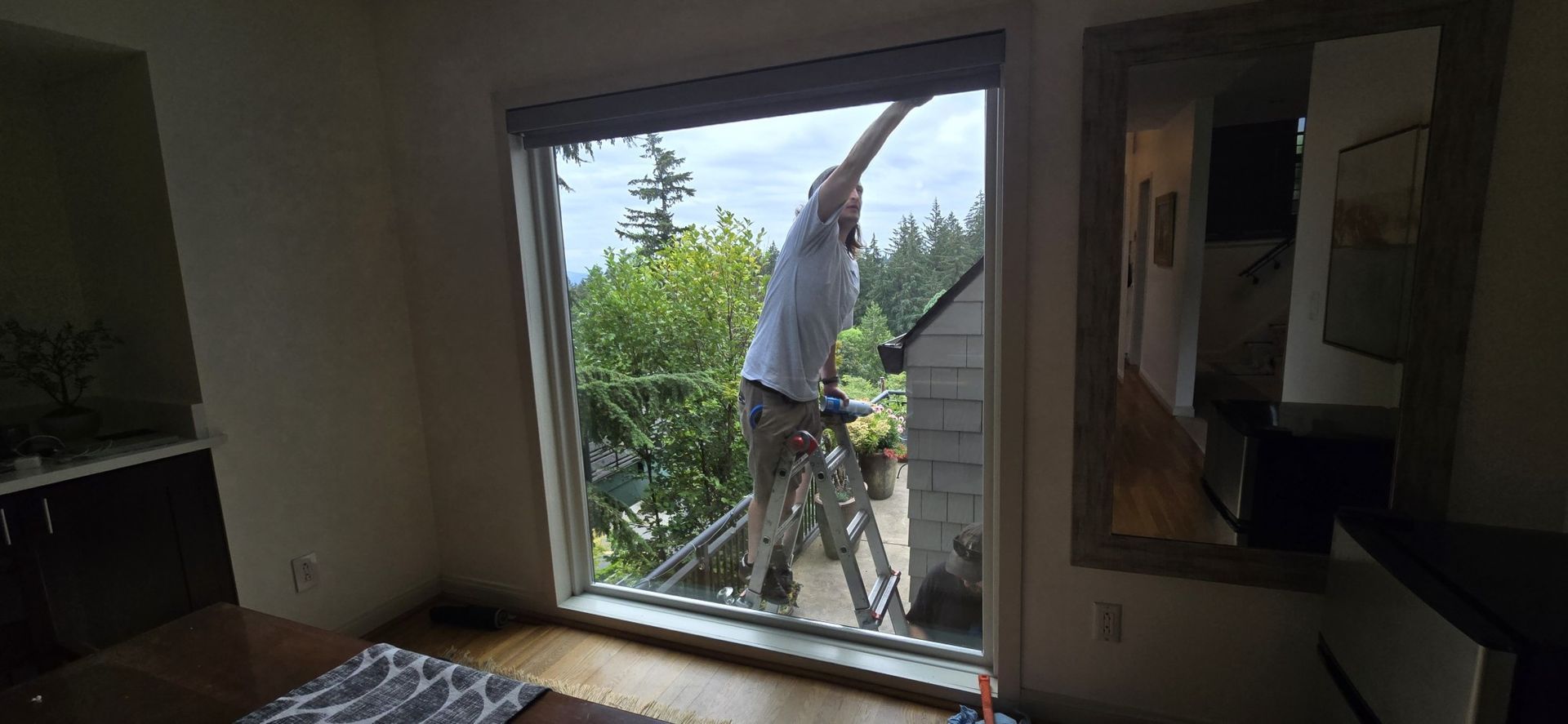 A man is standing on a ladder cleaning a window in a living room.