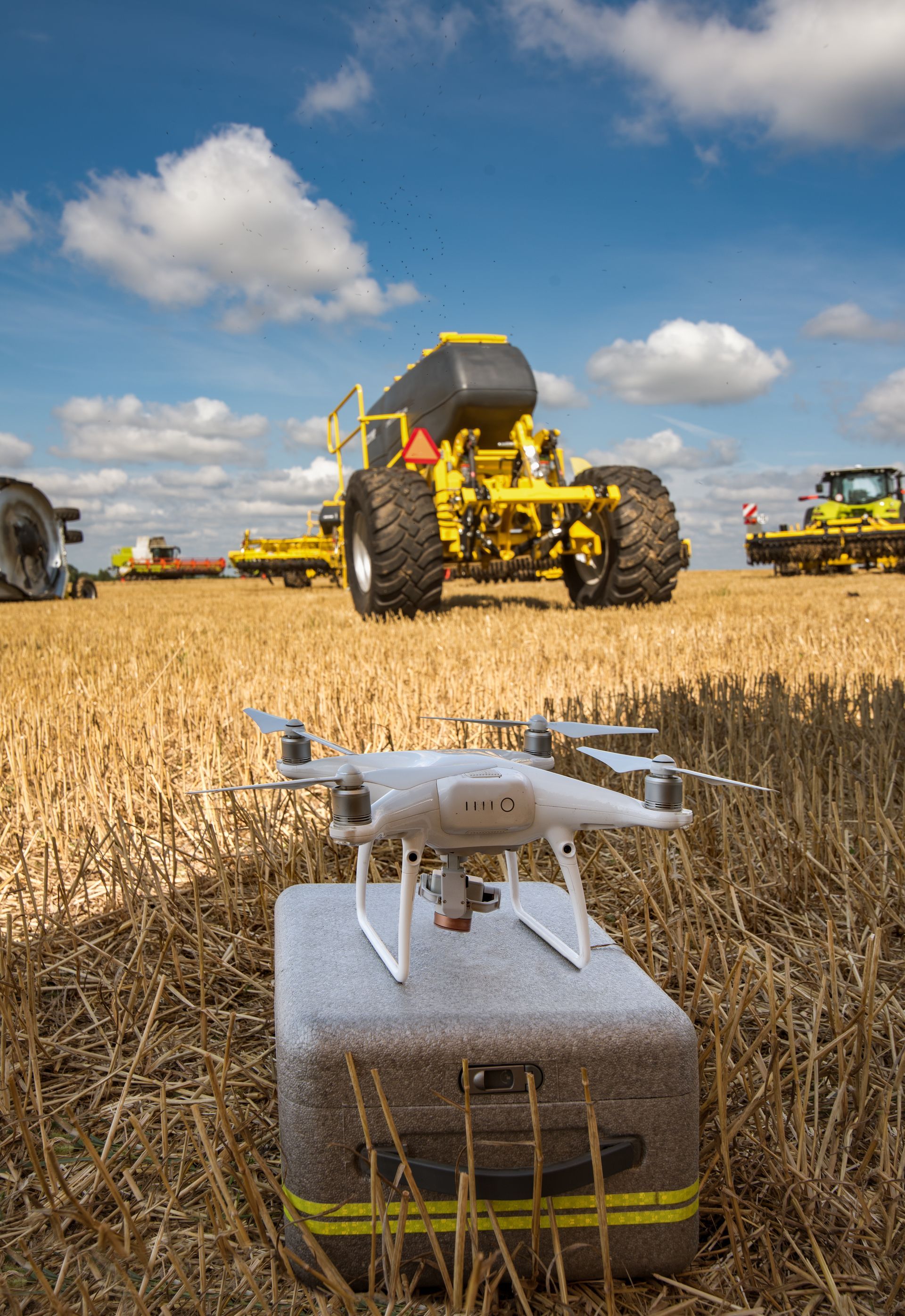Drone on case in harvested field, yellow farm machinery in background, under a blue sky.
