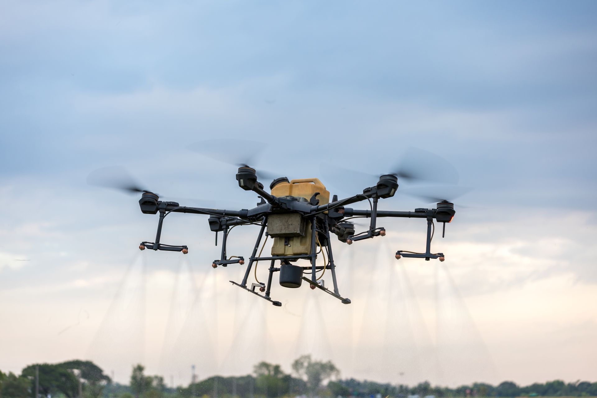 Agricultural drone spraying liquid over a field, with cloudy sky in the background.
