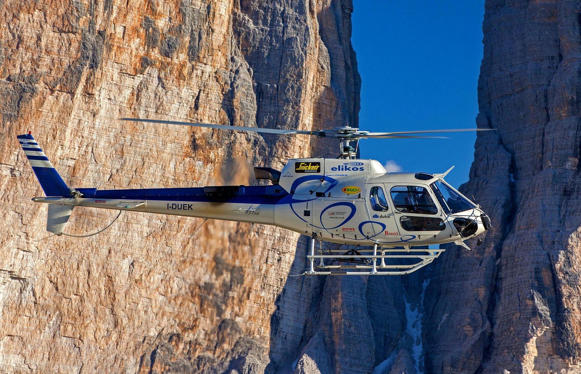 Helicopter flying near large rocky mountains; blue and white color scheme.
