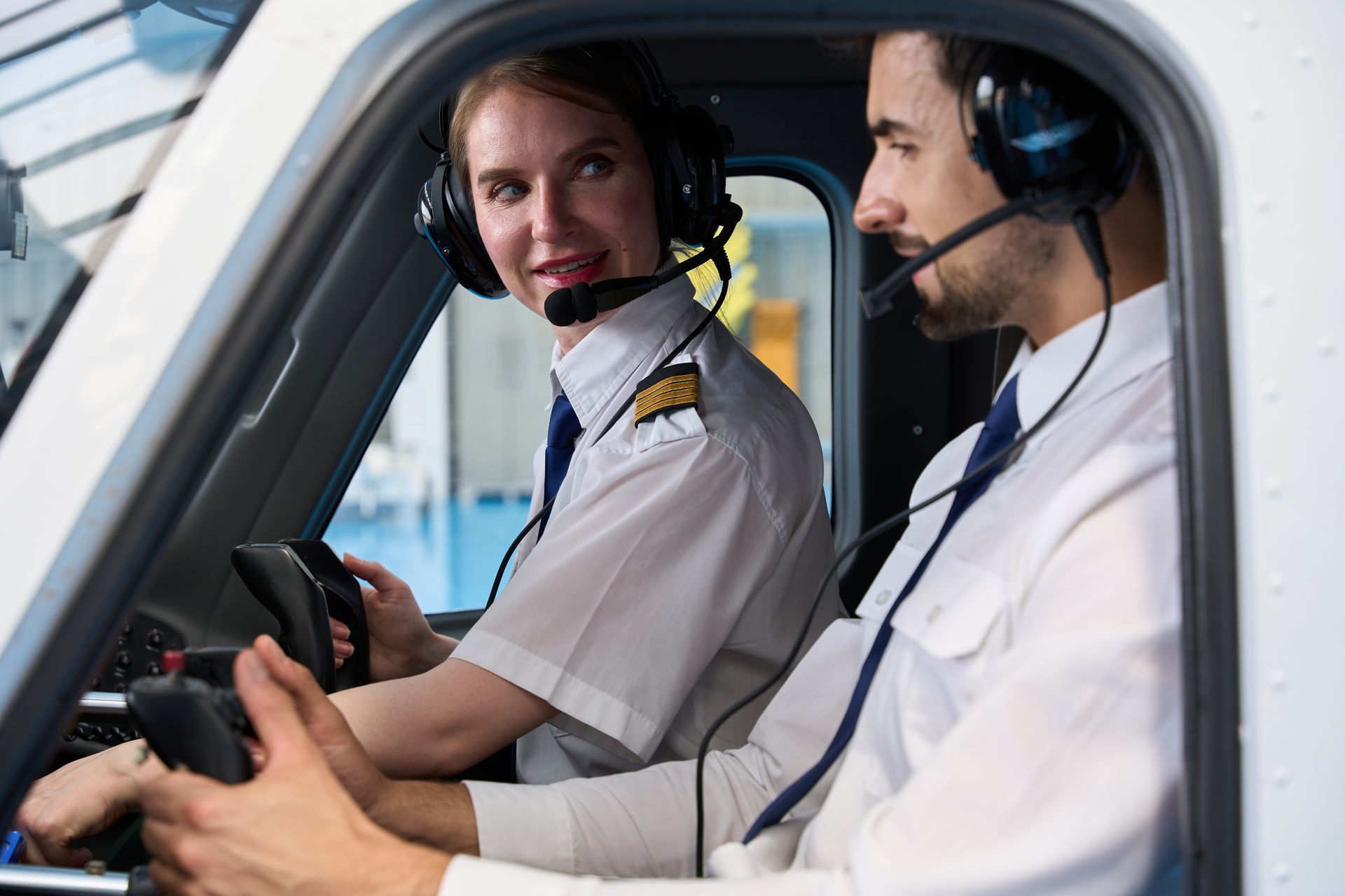 Two pilots in the cockpit, wearing headsets. The woman looks at the viewer, the man looks forward.