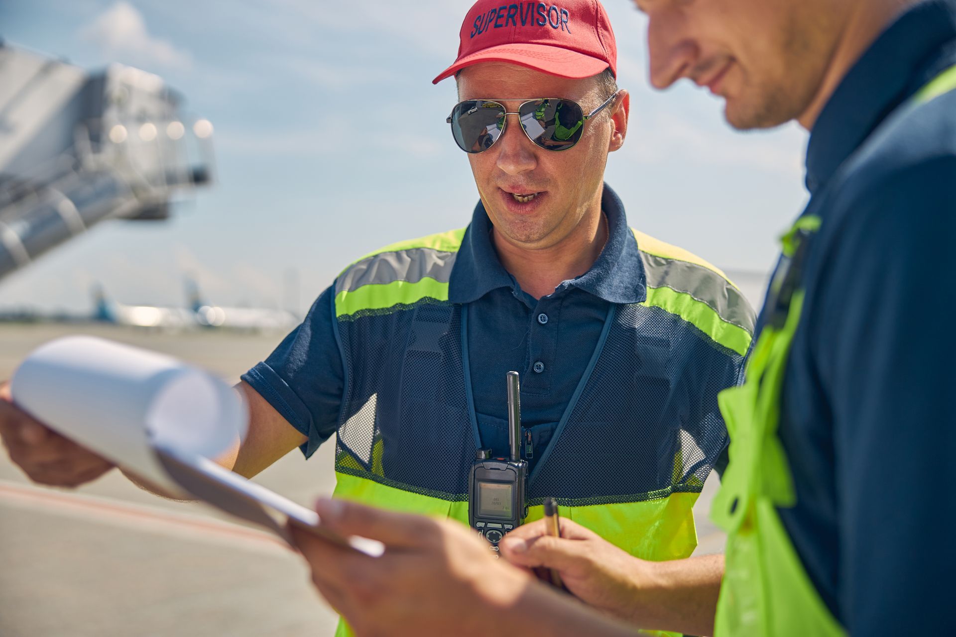 Two airport workers reviewing paperwork near a plane on the tarmac. One holds a radio.