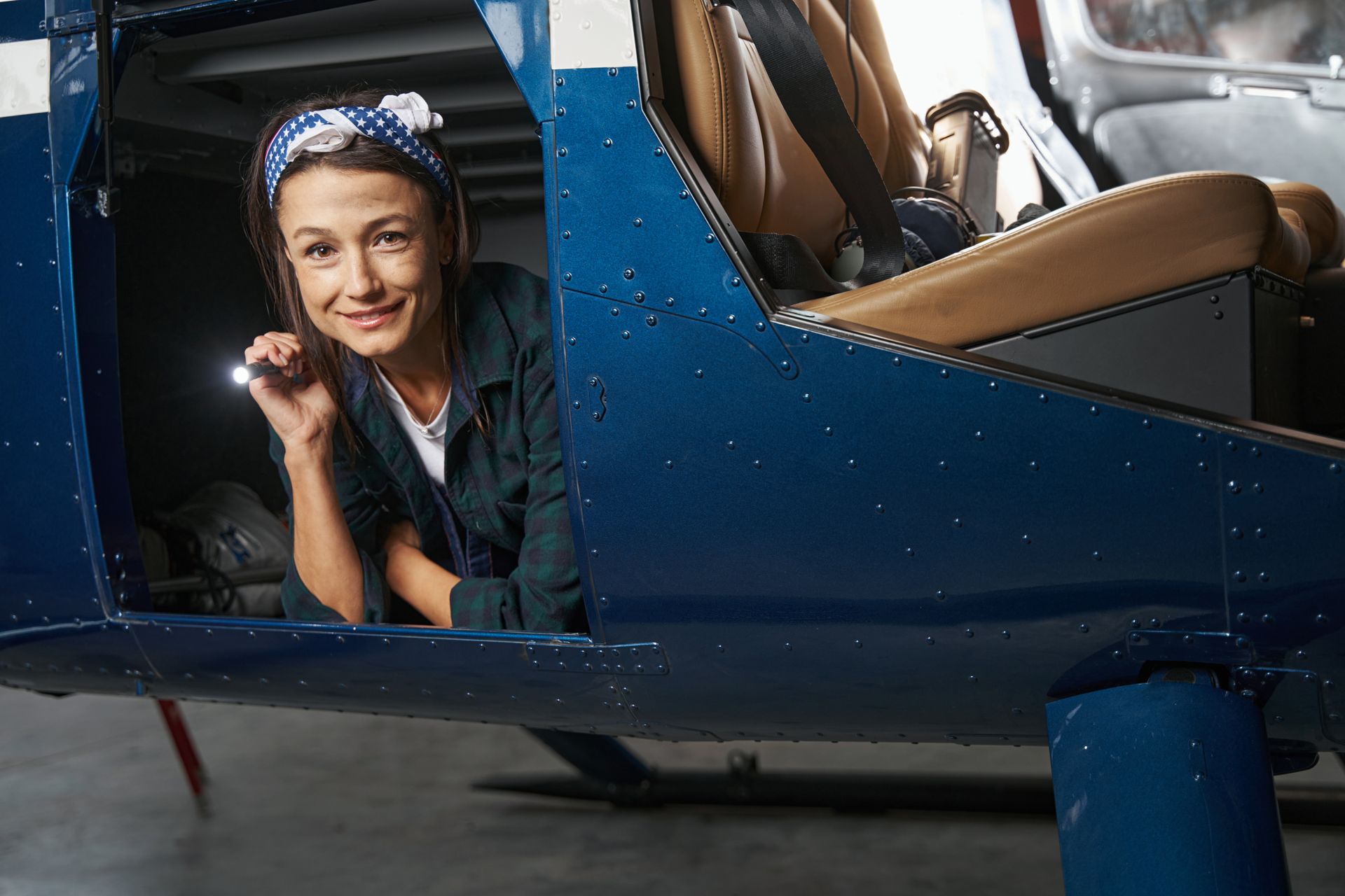 Woman with a headscarf, holding a flashlight in an open blue helicopter door, smiling.