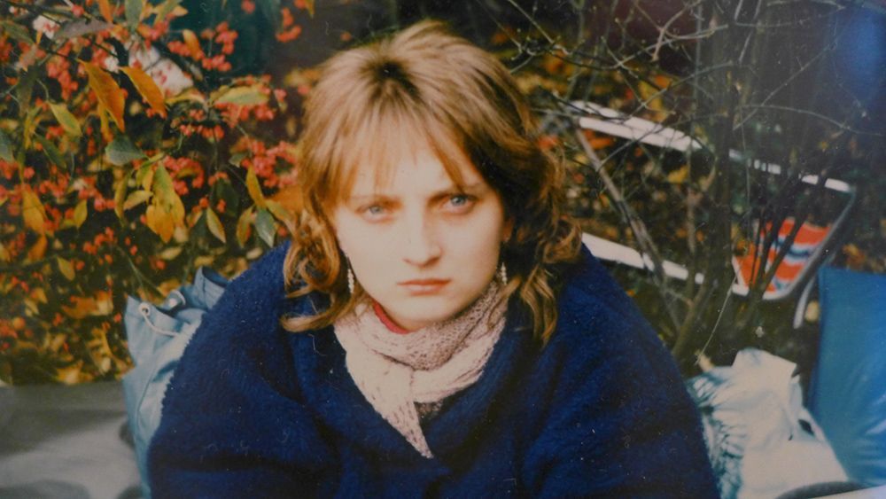 Person in a blue coat and scarf, looking at the camera against autumn foliage.