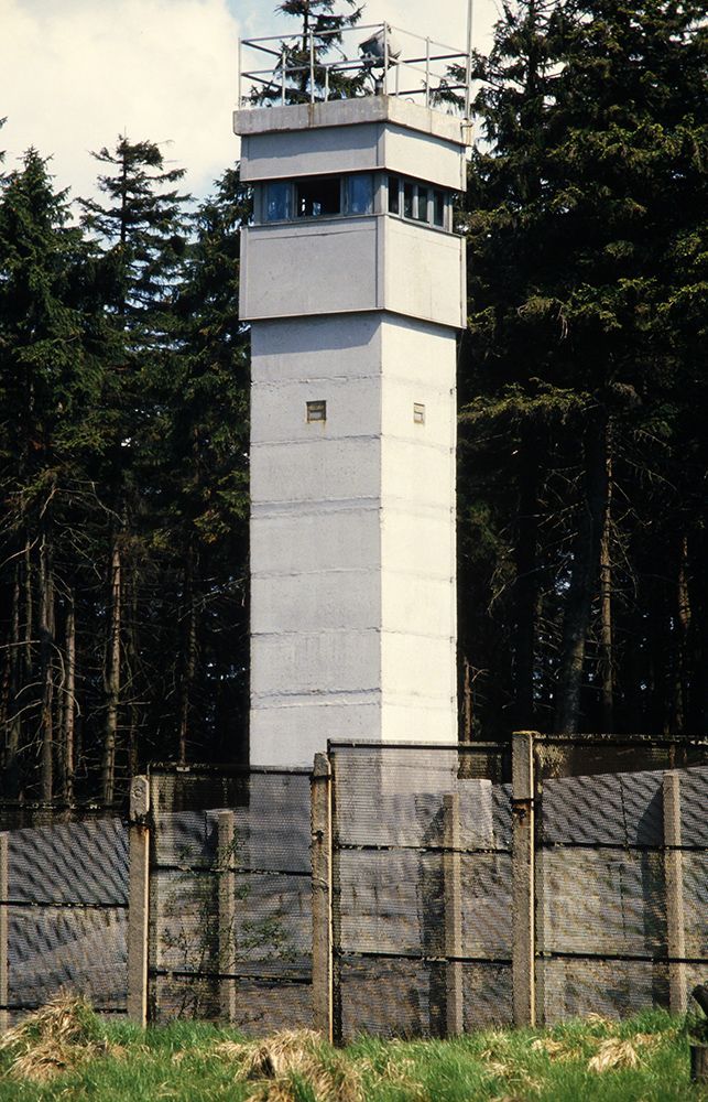 Tall white watchtower behind a wire fence, surrounded by trees.