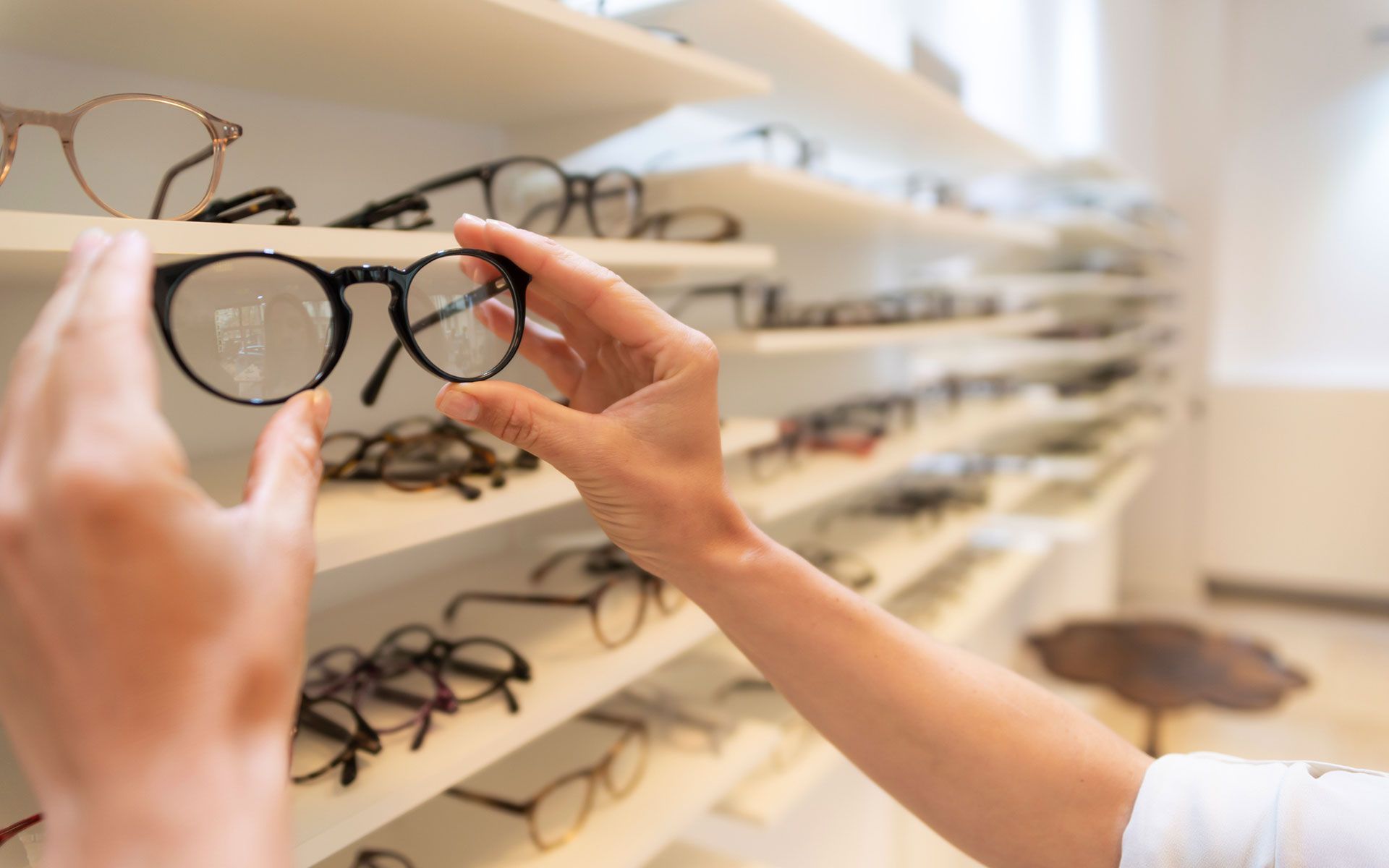 A person's hands picking a pair of round, black glasses from a display in an optical shop.