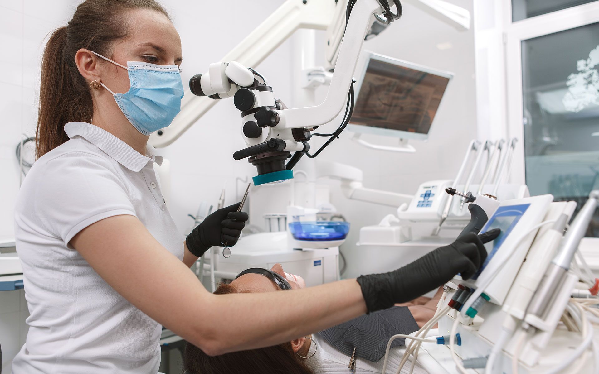 Dentist in mask and gloves operating dental equipment near patient in clinic.