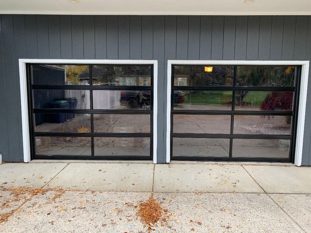 Two modern garage doors with black frames and glass panels. Set in a gray building with a concrete driveway.