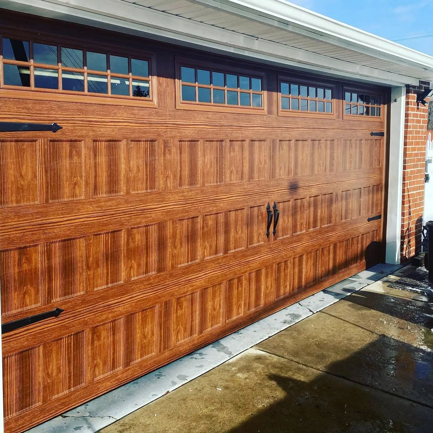 Wooden garage door with window panes and black handles beside a brick wall, with a wet concrete driveway
