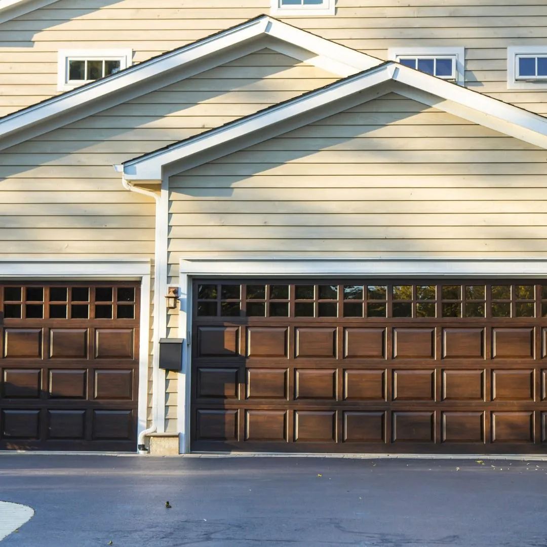 Two dark brown garage doors on a beige suburban house with a peaked roof.