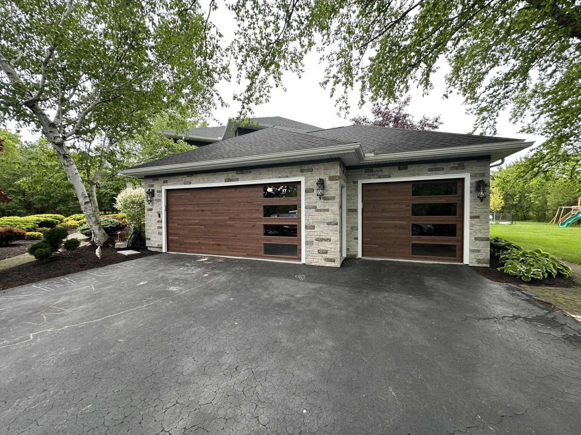 Suburban house with two garage doors, brick exterior, and a paved driveway under leafy trees