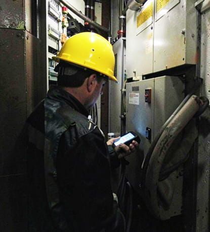 Un trabajador con casco amarillo utiliza un teléfono inteligente en una estrecha sala de equipos industriales.