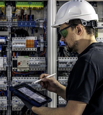 Técnico con casco y gafas de seguridad utilizando una tableta junto a paneles de control eléctrico.