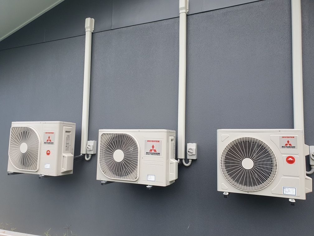 Three wall-mounted Mitsubishi air conditioning units with white piping on a gray building.— Cool Tropics Air Conditioning & Refrigeration in Mourilyan Harbour, QLD