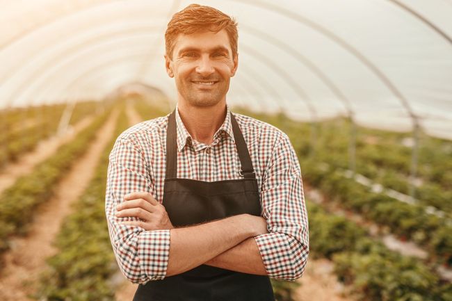 A man in an apron is standing in a greenhouse with his arms crossed.