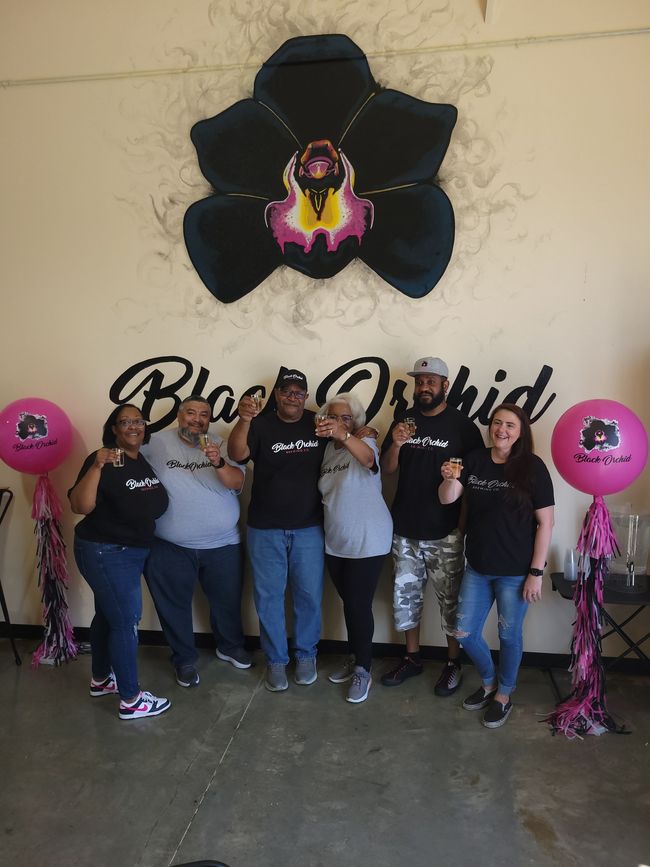 Group posing beneath a giant black flower mural, flanked by pink balloons and a Black Orchid sign.